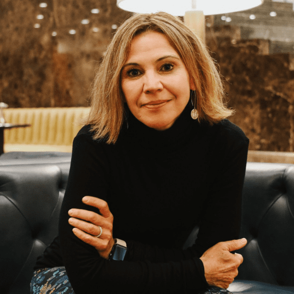 A woman with shoulder-length blonde hair, wearing a black turtleneck and earrings, sits on a blue sofa with her arms crossed in a warmly lit indoor setting.