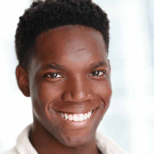 A young man with short curly hair smiles at the camera. He is wearing a light-colored shirt and stands in front of a softly blurred background.