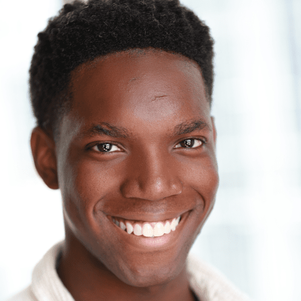 A young man with short curly hair smiles at the camera. He is wearing a light-colored shirt and stands in front of a softly blurred background.