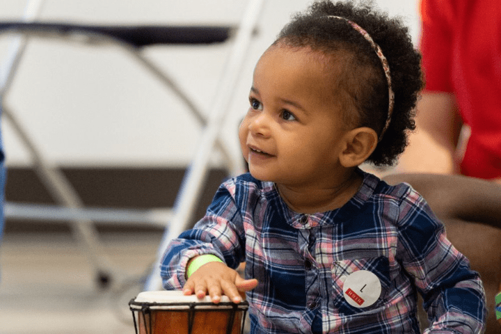 A young child wearing a plaid shirt and headband smiles while playing a small drum indoors.