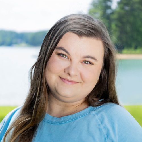 Woman with long brown hair wearing a light blue shirt, smiling outdoors with a lake and trees in the background.