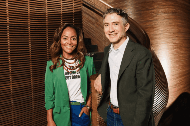 Two people stand in front of a curved wooden staircase, both smiling. One wears a green blazer and the other a dark jacket over a light shirt.