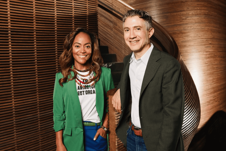 Two people stand in front of a curved wooden staircase, both smiling. One wears a green blazer and the other a dark jacket over a light shirt.