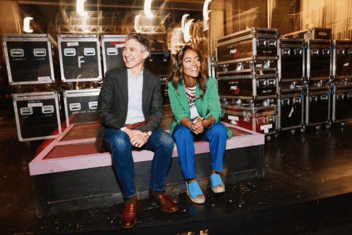 Two people sit and smile on a stage platform, surrounded by equipment cases and lighting, appearing relaxed in a backstage area.