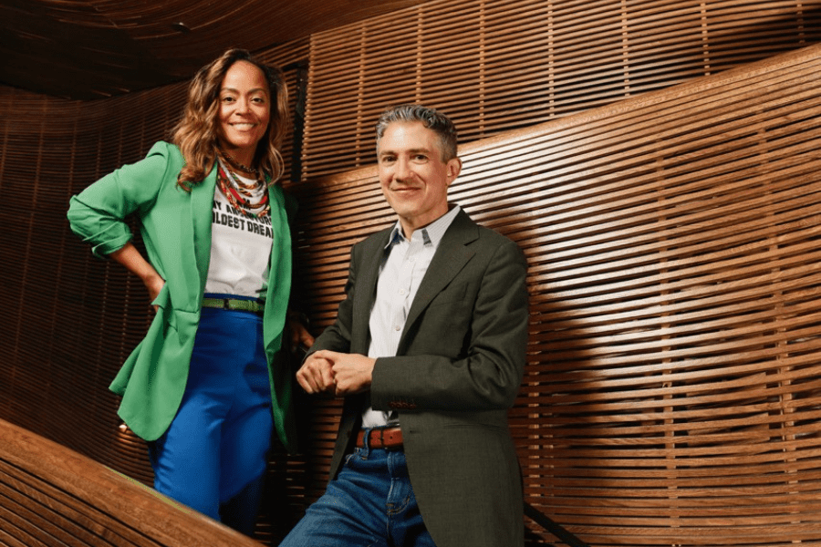 Two people stand on a wooden staircase, both wearing blazers; the woman on the left wears a green blazer and blue pants, the man on the right wears a gray blazer and jeans.