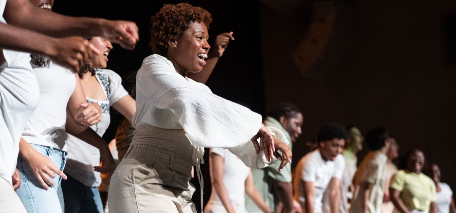 A group of people stands in a row on stage, energetically dancing and smiling. The lighting highlights the central figure in white clothing.