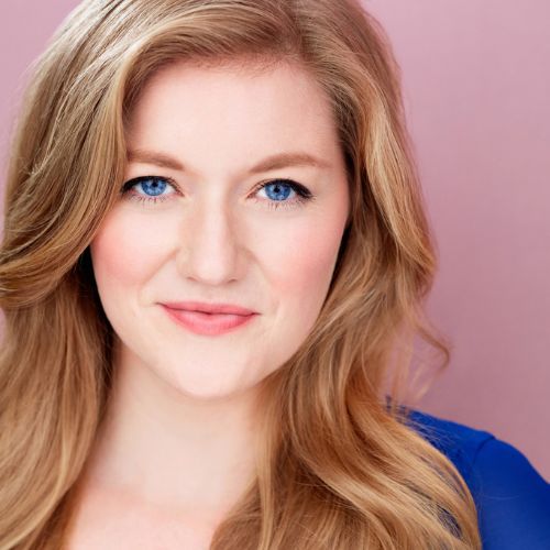 A woman with long blonde hair and blue eyes smiles slightly, wearing a blue top, against a pink background.