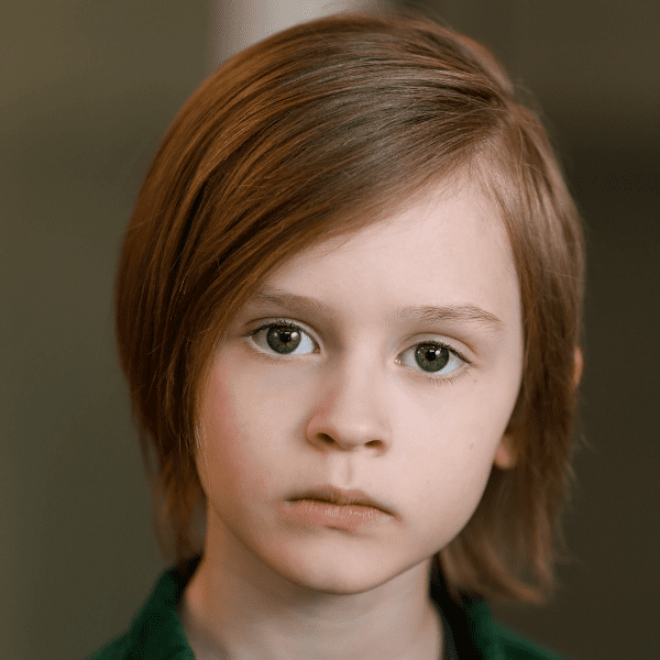 A young child with straight, shoulder-length light brown hair and light skin looks directly at the camera with a neutral expression. The background is out of focus.