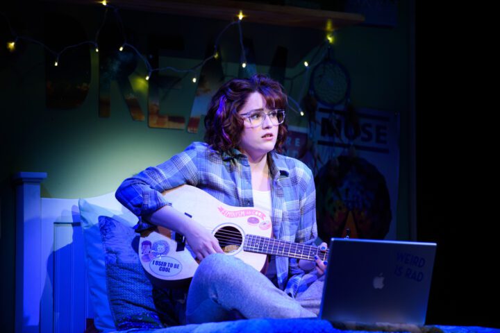 A person in glasses sits on a bed playing guitar, looking concerned. A laptop and string lights are visible in the cozy, decorated bedroom.