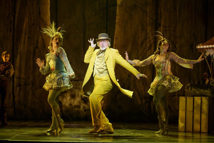 A person in a yellow suit and hat dances on stage with two performers in elaborate green costumes and headdresses, set against a forest-themed backdrop.