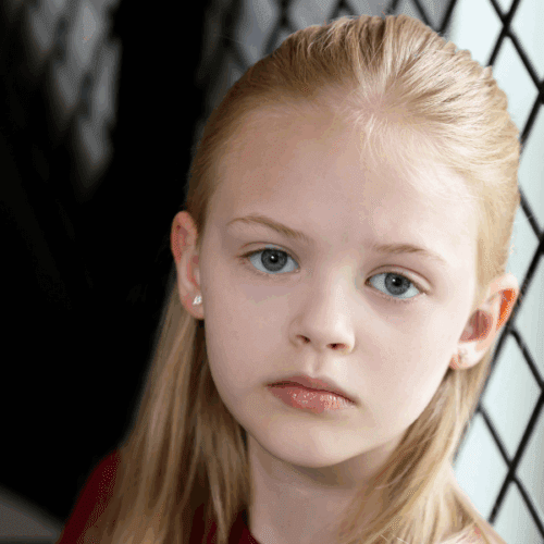 A young girl with long blonde hair and blue eyes looks at the camera with a neutral expression, standing near a wire fence.