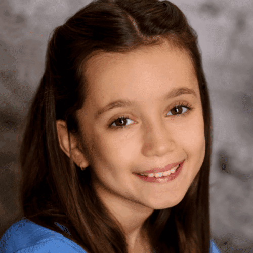 Smiling young girl with long brown hair wearing a blue top, posing in front of a gray textured background.