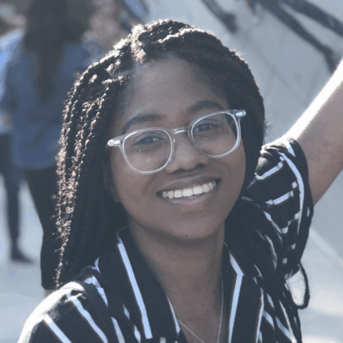 A person with braided hair and glasses smiles outdoors, wearing a black and white striped shirt. Other people are visible in the blurred background.