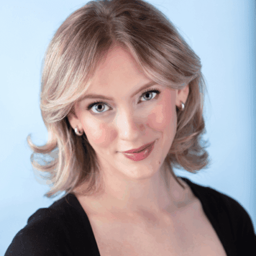 A woman with light blonde hair, light skin, and green eyes smiles softly at the camera. She is wearing a black top and small hoop earrings, with a light blue background.