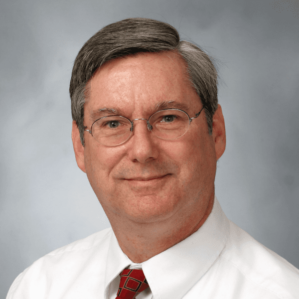 A middle-aged man with short gray hair, glasses, and a white shirt with a red patterned tie, posing in front of a neutral gray background.