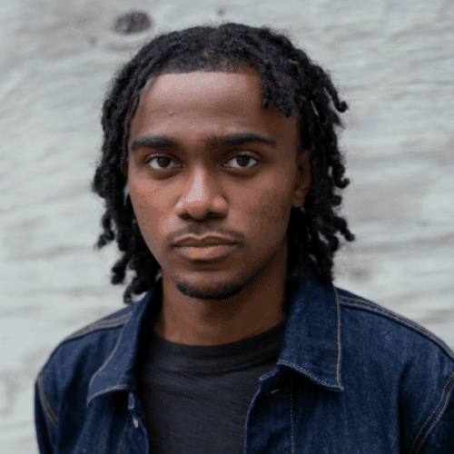 A young man with short dreadlocks and a denim jacket looks directly at the camera against a light-colored, textured background.