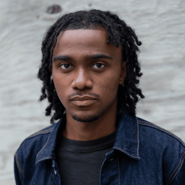 A young man with short dreadlocks and a denim jacket looks directly at the camera against a light-colored, textured background.
