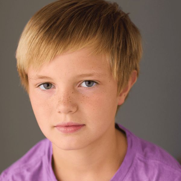 A young person with short blond hair and freckles, wearing a purple shirt, looks at the camera against a plain gray background.