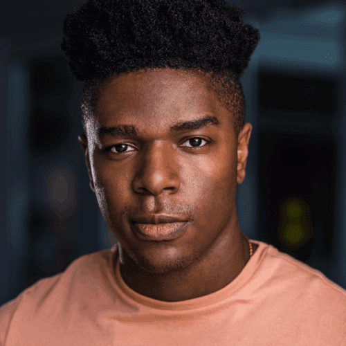 A young man with short curly hair wearing a peach-colored shirt looks directly at the camera with a neutral expression.