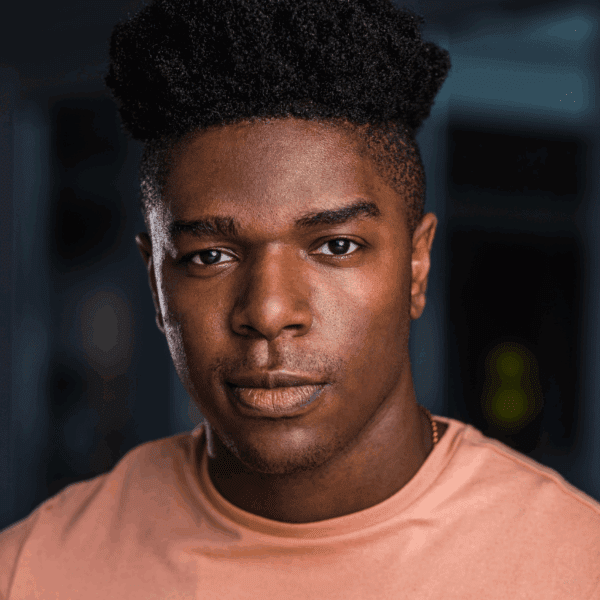 A young man with short curly hair wearing a peach-colored shirt looks directly at the camera with a neutral expression.