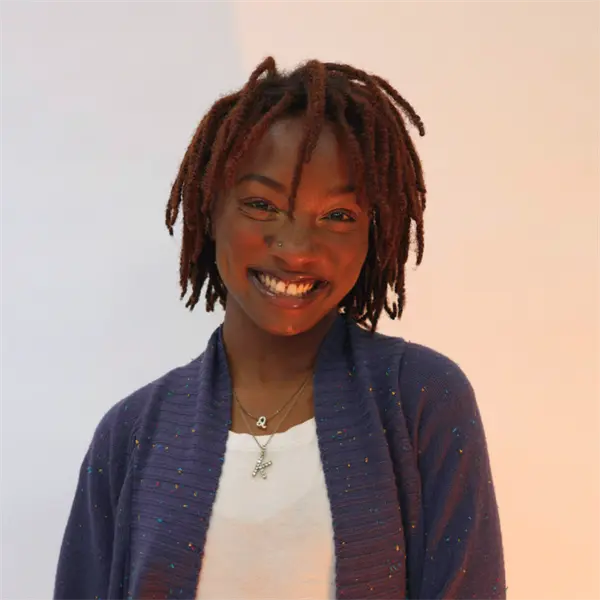 A person with short, twisted dreadlocks smiles at the camera, wearing a blue cardigan over a white shirt and two necklaces, against a plain light background.