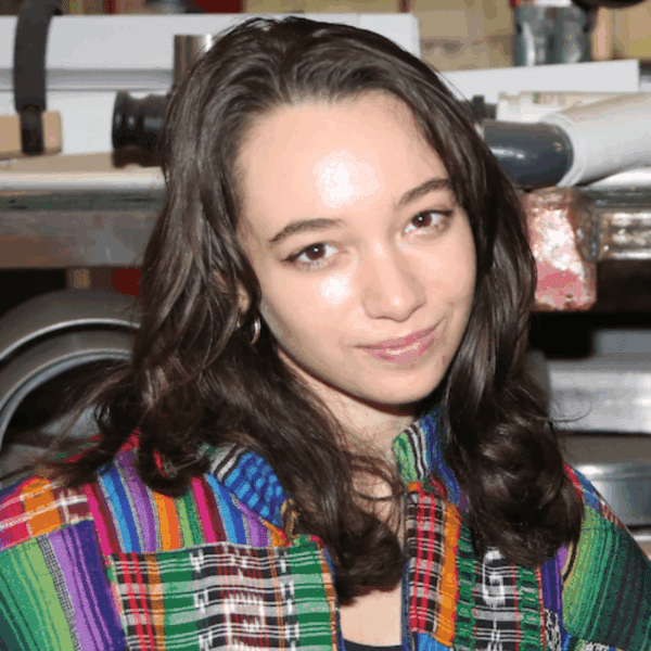 A person with long brown hair wearing a colorful patterned shirt stands indoors, looking at the camera with a slight smile.