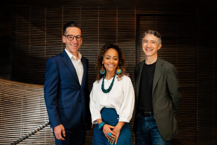 Brandon Kahn, Tinashe Kajese-Bolden, and Christopher Moses indoors in front of a wooden slatted wall. One man is in a blue suit, a woman is in a white blouse and blue pants, and another man is in a dark blazer and black shirt.