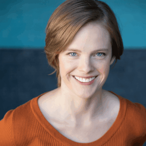 A person with short light brown hair, wearing an orange top, is smiling at the camera with a blue and dark background.