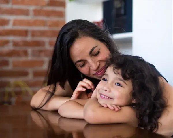 A woman and a young girl with curly hair smile while resting their arms on a wooden table, with a brick wall and shelves in the background.