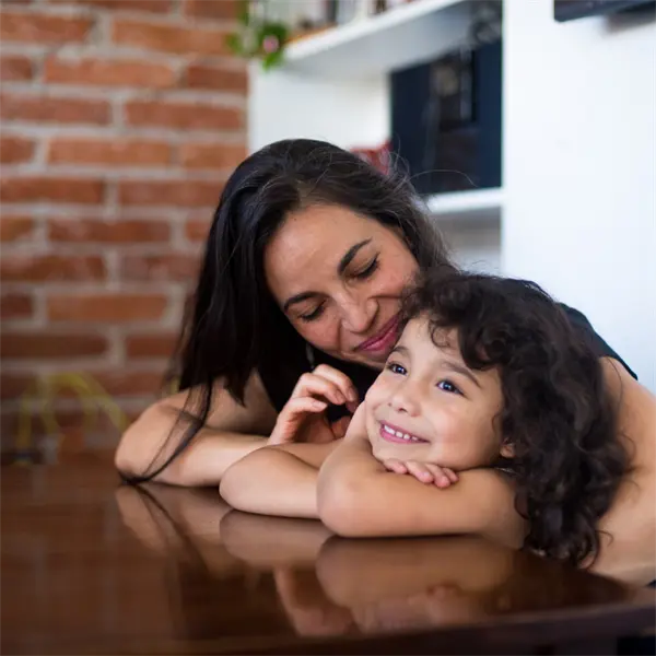 A woman and a young girl with curly hair smile while resting their arms on a wooden table, with a brick wall and shelves in the background.