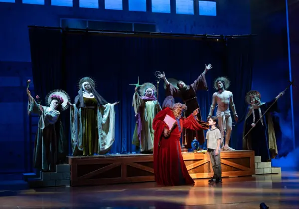 A stage scene features actors dressed as religious figures with halos, while a robed figure gestures toward a boy standing at the front. Blue lighting creates a dramatic atmosphere.