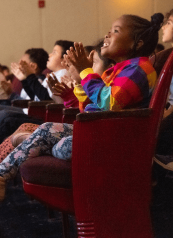 Children sit in theater seats, clapping and watching something off-camera. One child in a colorful jacket is smiling and appears particularly engaged.