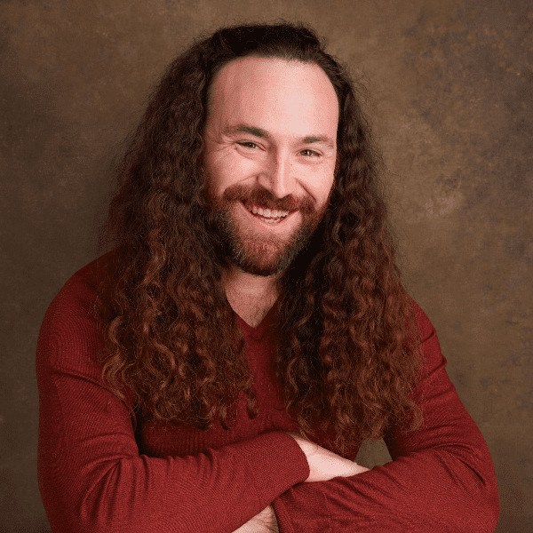 A person with long curly brown hair and a beard, wearing a red sweater, smiles with arms crossed against a brown textured background.