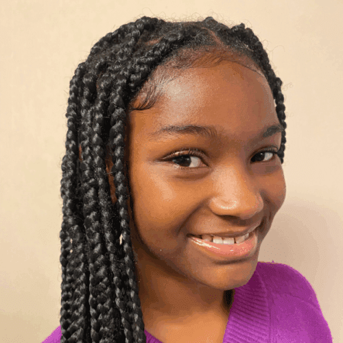 Smiling young girl with braided hair, wearing a purple top, shown from the shoulders up against a plain background.