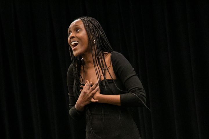 Woman with long braids wearing a black outfit stands in front of a black curtain, holding her hands to her chest and speaking with an expressive facial expression.