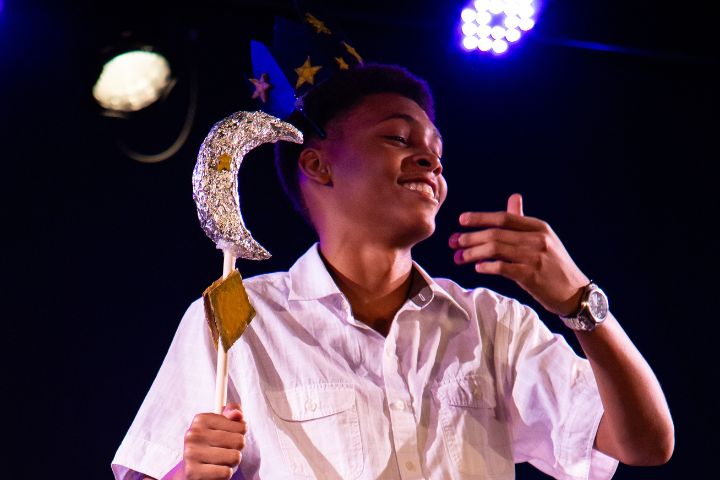 A person in a white shirt holds a crescent moon staff and wears a star-covered wizard hat, standing under stage lights.