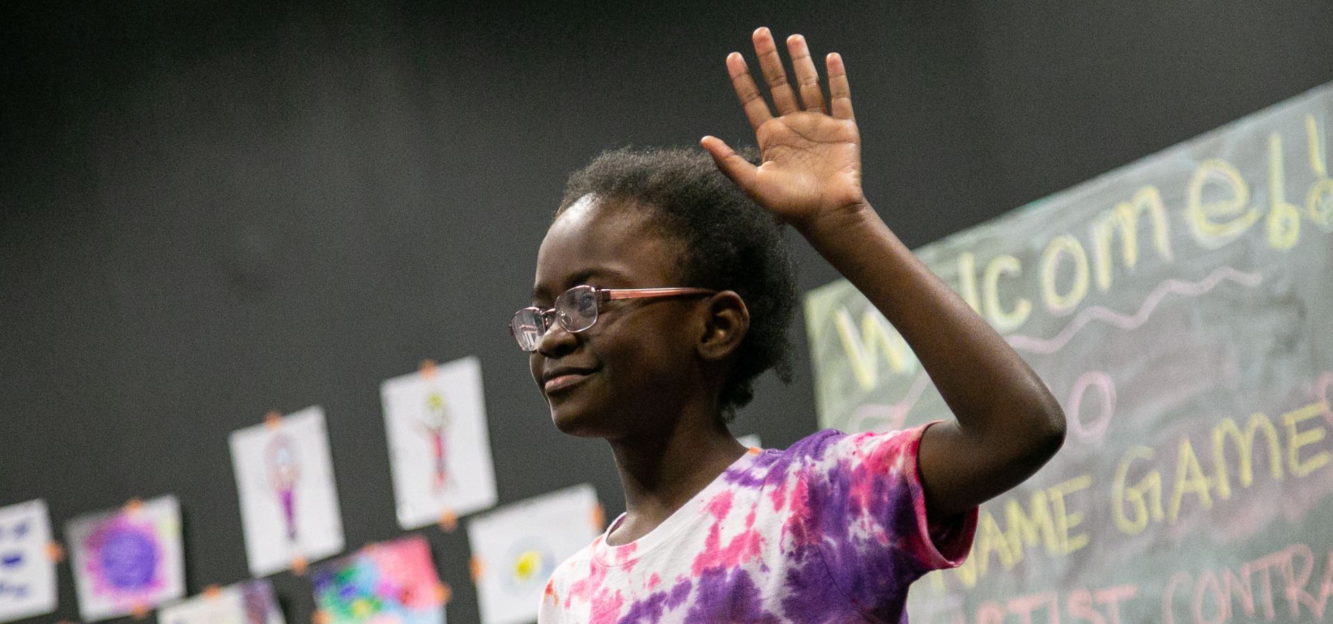 A child wearing glasses and a tie-dye shirt raises their hand in a classroom with drawings on the wall and a chalkboard in the background.