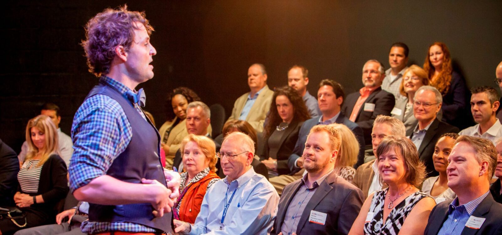 A man speaks to an audience seated in rows, with most people attentively watching him in a well-lit indoor setting.