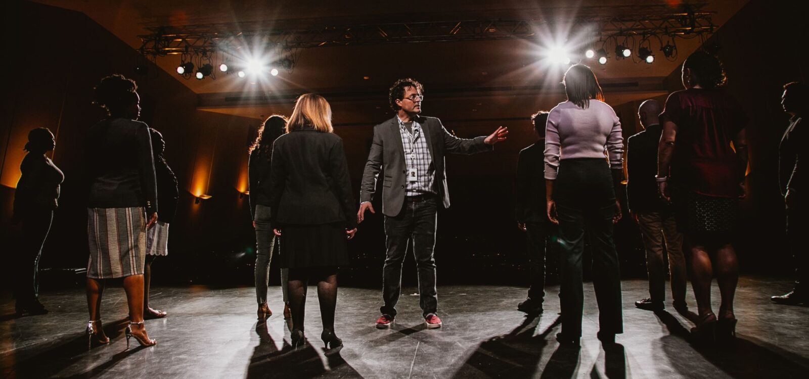 A group of people stands on a stage under bright lights, with one person in the center gesturing as if addressing the others.