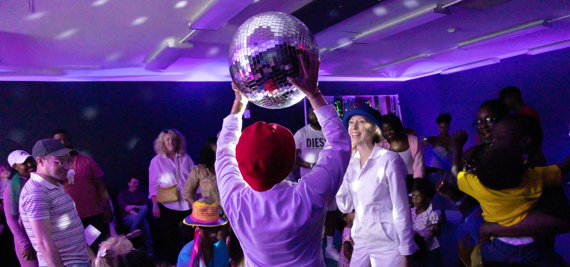A person holding up a disco ball in a crowded, dimly lit room with purple lighting as people watch and interact.