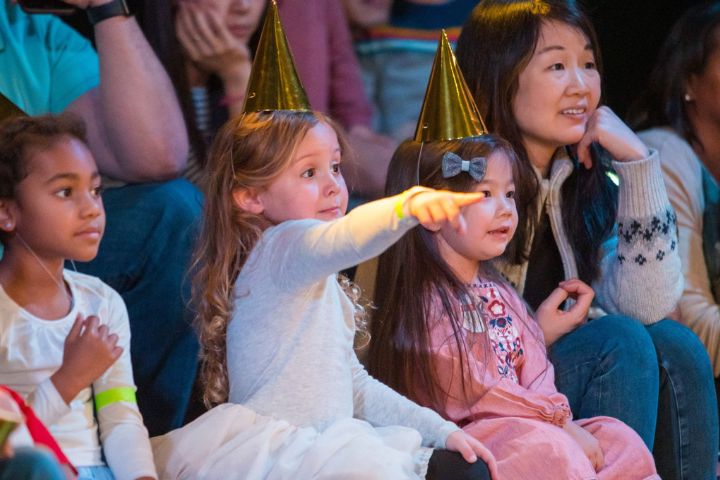 A group of children and an adult sit closely together, watching something off-camera; two children wear gold party hats, and one is pointing excitedly.