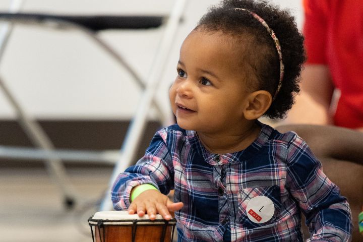 Brave + Kind Story Time A young child in a plaid shirt plays a small drum, smiling and looking to the side. The child wears a headband and a name tag on their shirt.