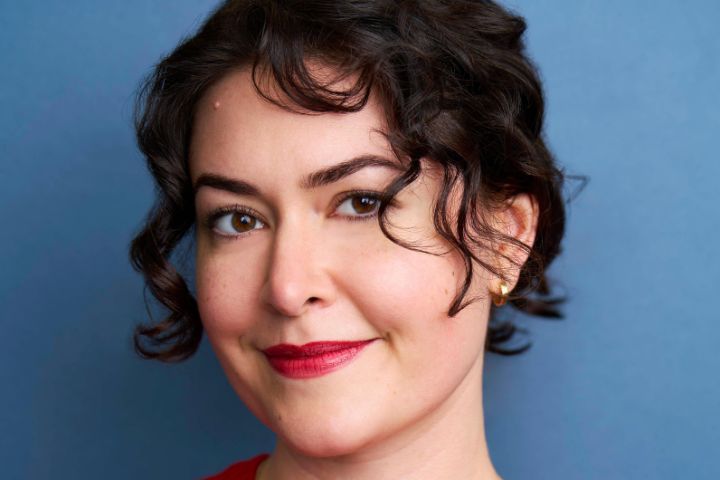 A person with short, curly dark hair and red lipstick smiles softly at the camera against a plain blue background.