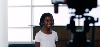A young girl wearing a white shirt stands in front of a window and smiles while being filmed by a camera.