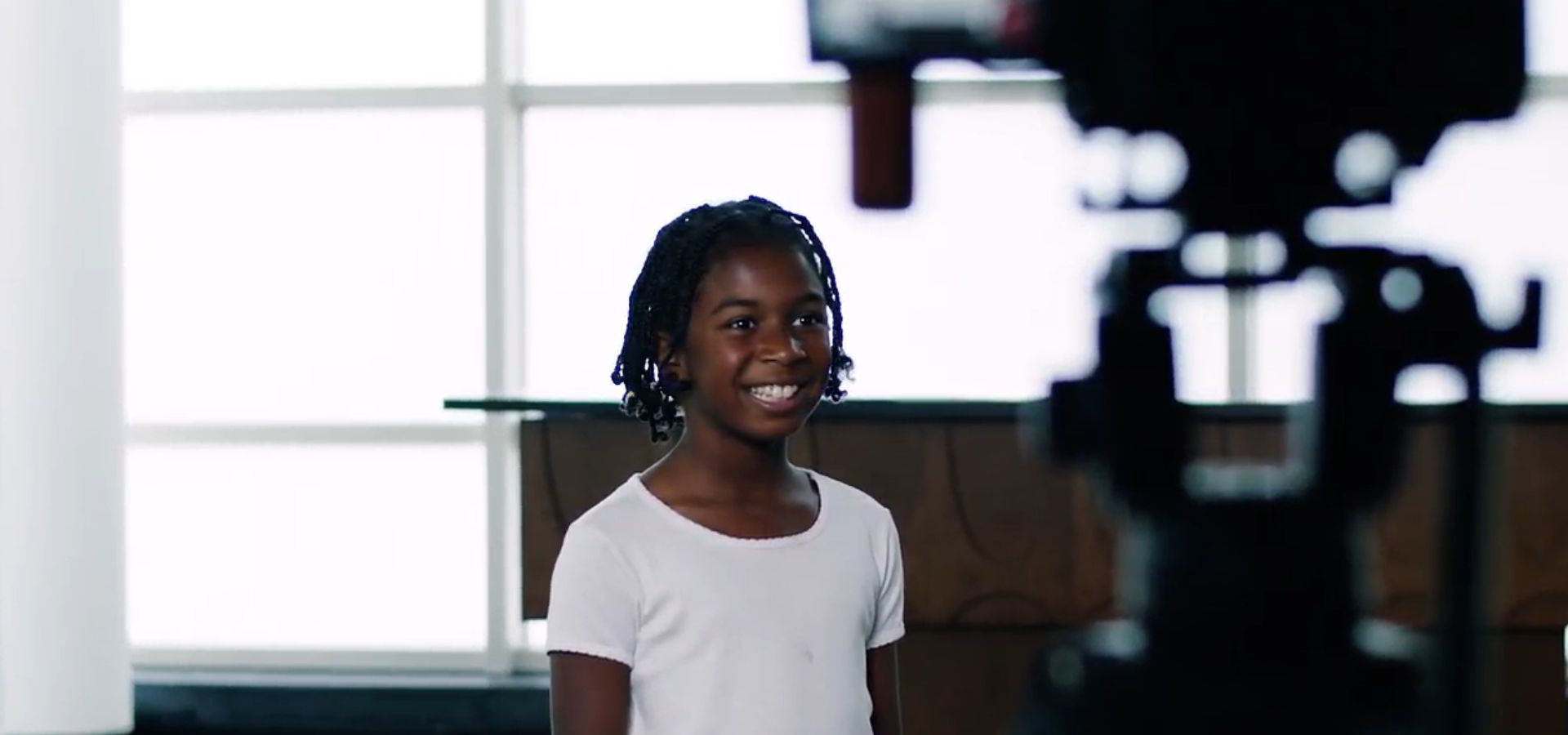 A young girl wearing a white shirt stands in front of a window and smiles while being filmed by a camera.