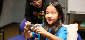 A young girl in a blue shirt holds a digital camera, taking a photo, while another person stands behind her watching.