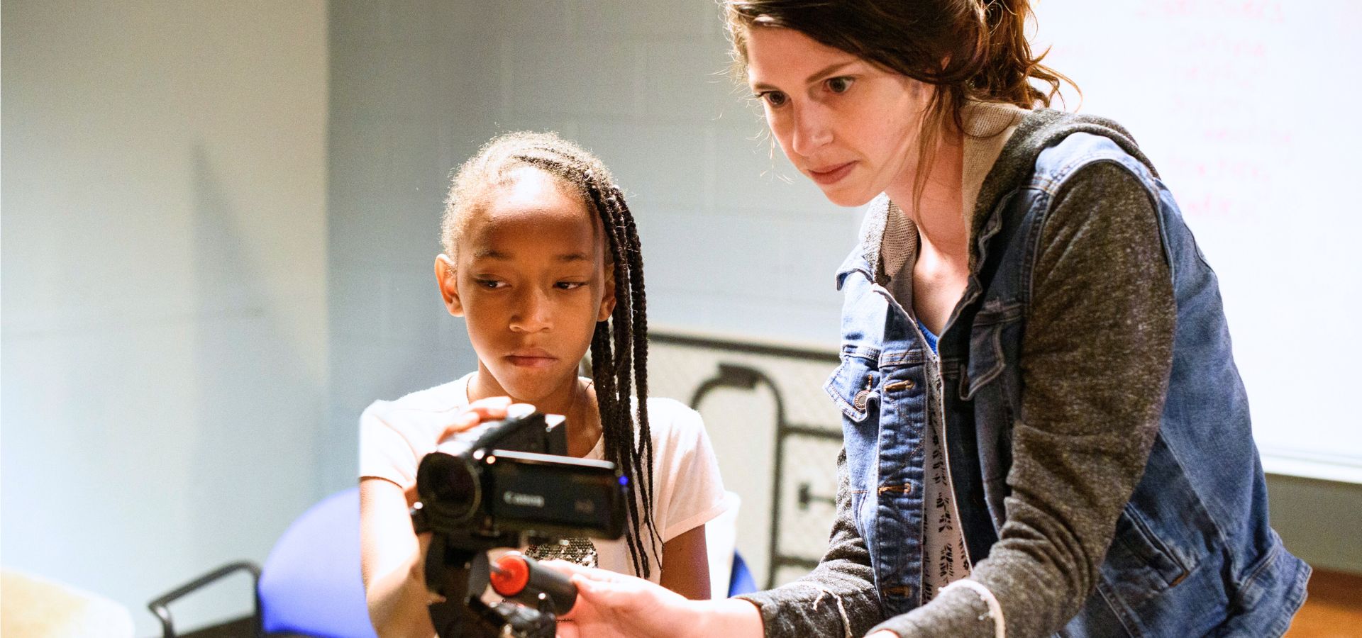 An adult and a child operate a video camera together in an indoor setting, with the adult guiding the child’s hands on the camera controls.