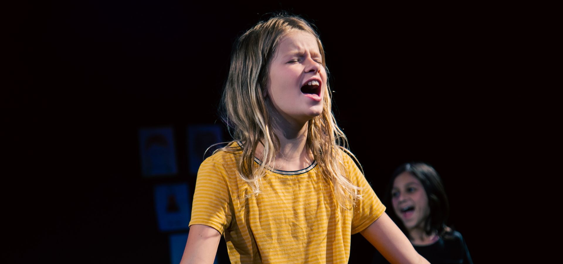 A young girl in a yellow shirt stands with arms outstretched, singing or speaking passionately on stage; another child is visible in the background.