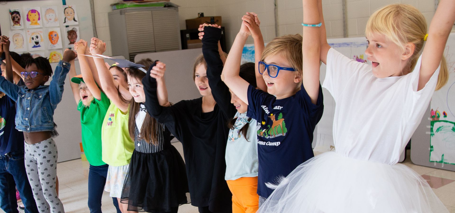 A group of young children stand in a row indoors, holding hands and raising their arms, smiling and appearing to celebrate or perform together.