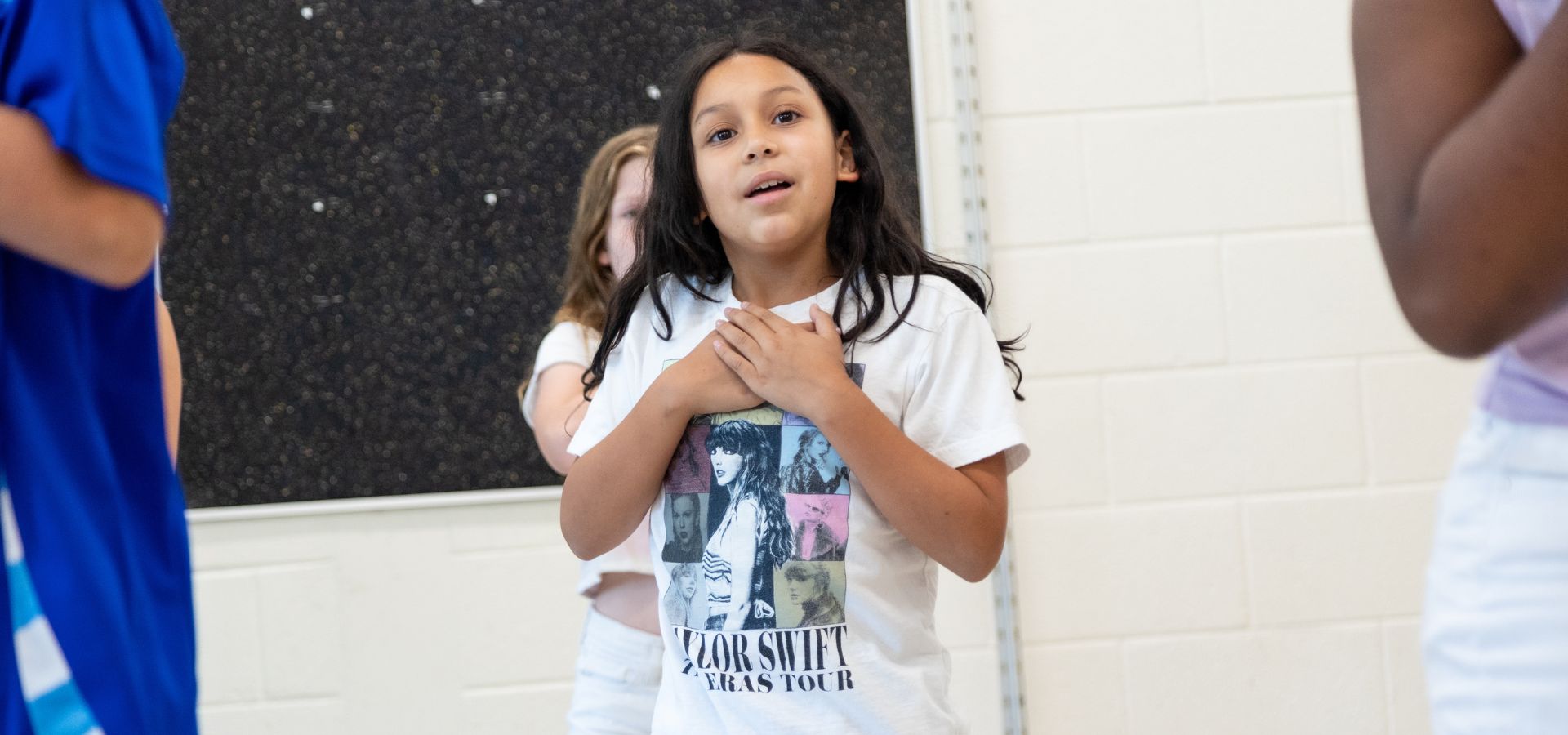 A young girl stands indoors with hands clasped to her chest, wearing a white Taylor Swift Eras Tour T-shirt. Other children are partially visible around her.